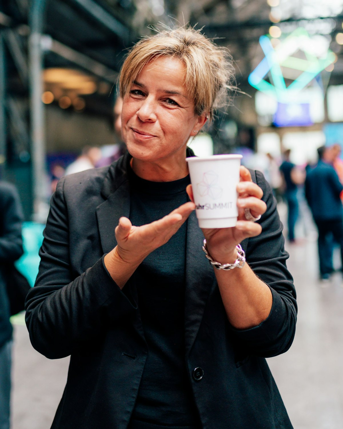 Eine lächelnde Frau in einem schwarzen Blazer hält einen weißen Pappbecher mit einem Logo in der Hand und gestikuliert, während sie in einem hellen Veranstaltungsraum mit Menschen und Lichtern im Hintergrund steht.