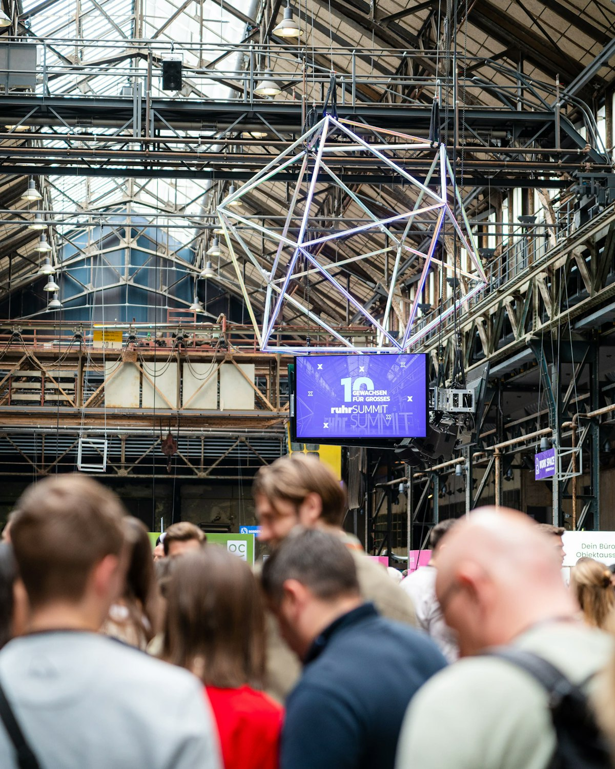 Eine Menschenmenge steht in einer Halle im Industriestil mit Metallträgern und Glasdecken. Über ihnen hängen eine geometrische Kunstinstallation und ein Bildschirm mit der Aufschrift "10th Anniversary Web Summit".