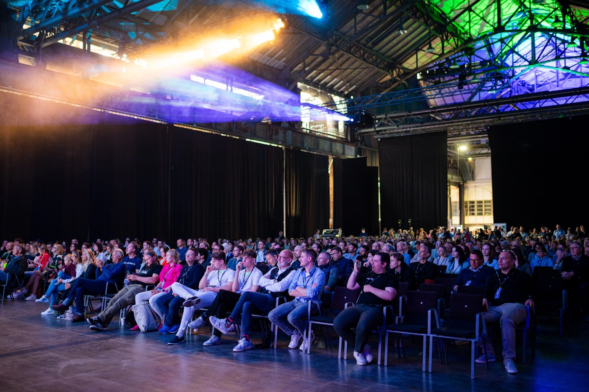 Ein großes Publikum sitzt in Stuhlreihen in einem geräumigen Saal im Industriestil mit hohen Decken und bunten Lichtern und verfolgt eine Veranstaltung oder Präsentation auf der Bühne.