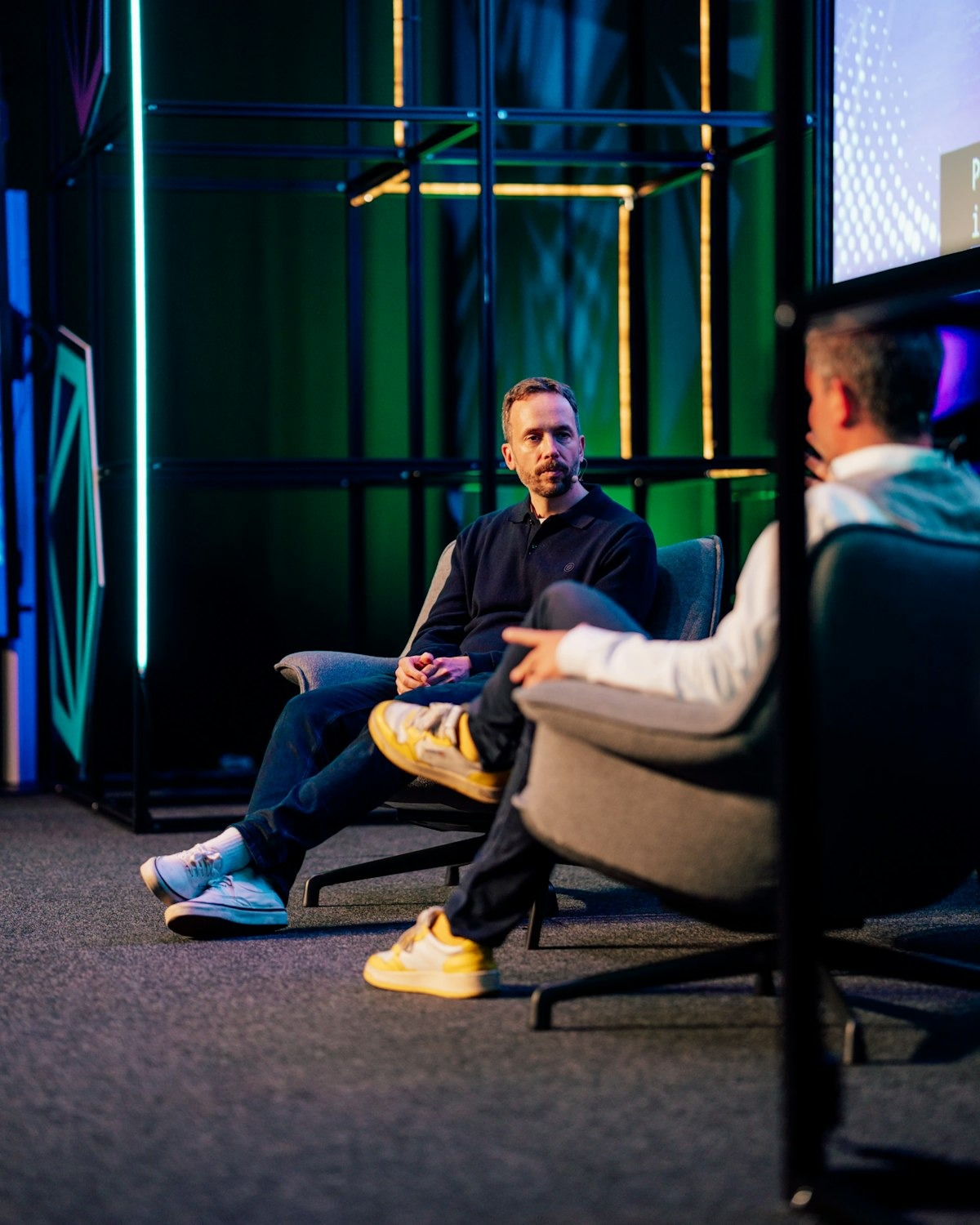 Zwei Personen sitzen auf grauen Stühlen auf einer Bühne und führen ein Gespräch. Die eine Person ist dem Publikum zugewandt, während die andere von hinten gesehen wird. Die Szene ist mit bunten vertikalen Lichtern und geometrischen Formen im Hintergrund beleuchtet.