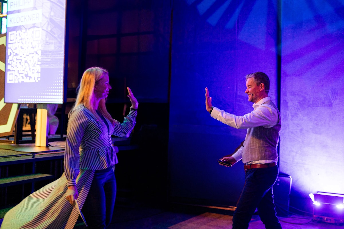 Eine Frau und ein Mann geben sich auf einer Bühne mit blauer und lila Beleuchtung ein High Five. Im Hintergrund ist ein großer Bildschirm mit einem QR-Code und Text zu sehen.
