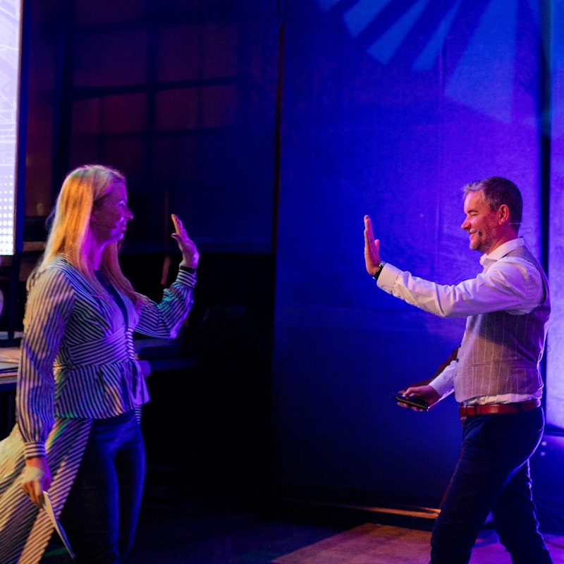 Eine Frau und ein Mann geben sich auf einer Bühne mit blauer und lila Beleuchtung ein High Five. Im Hintergrund ist ein großer Bildschirm mit einem QR-Code und Text zu sehen.