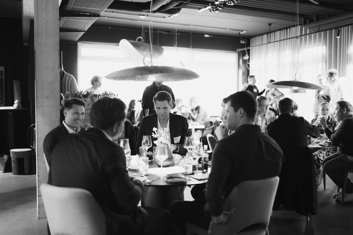 Eine Gruppe von formell gekleideten Personen sitzt um einen Tisch in einem Restaurant oder Bankett und unterhält sich. Die Szene ist lebhaft, im Hintergrund sind andere Gäste und das Personal zu sehen. Das Bild ist in schwarz-weiß gehalten.