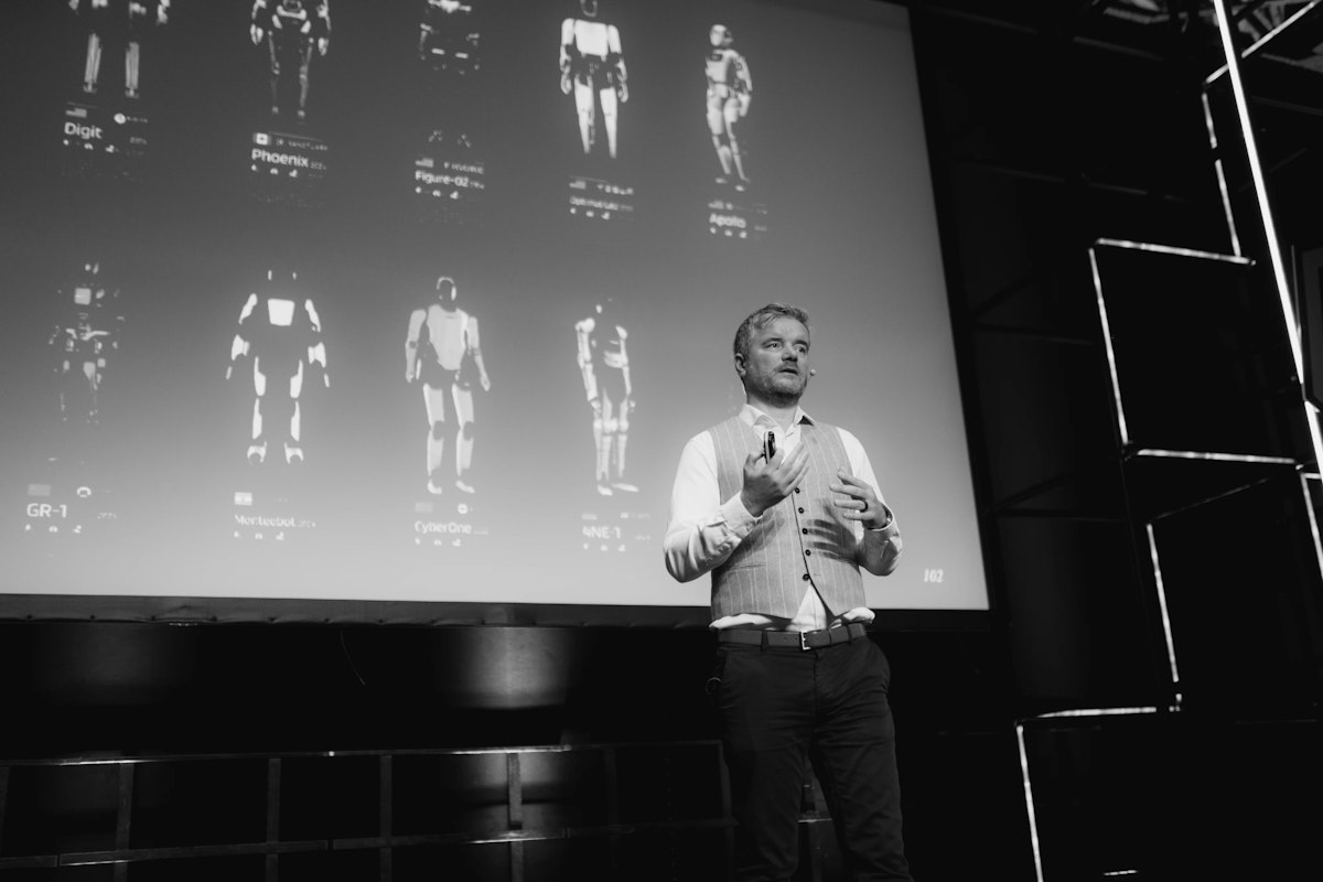 Ein Mann, der eine Weste und ein Headset trägt, steht auf der Bühne und spricht mit Gesten vor einem großen Bildschirm, auf dem Bilder von humanoiden Robotern zu sehen sind. Die Szene ist in schwarz-weiß gehalten.